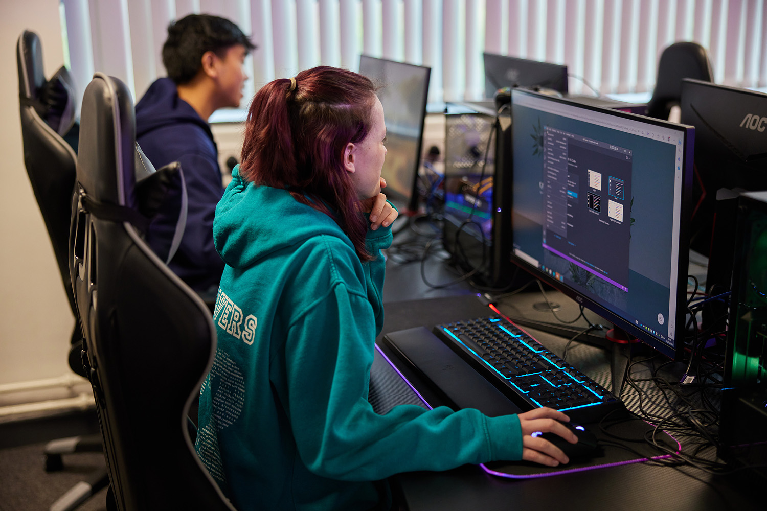 Two computing students sat next to one another, using high-tech computing equipment. They are sat on gaming chairs and look enthusiastic about what is on screen.
